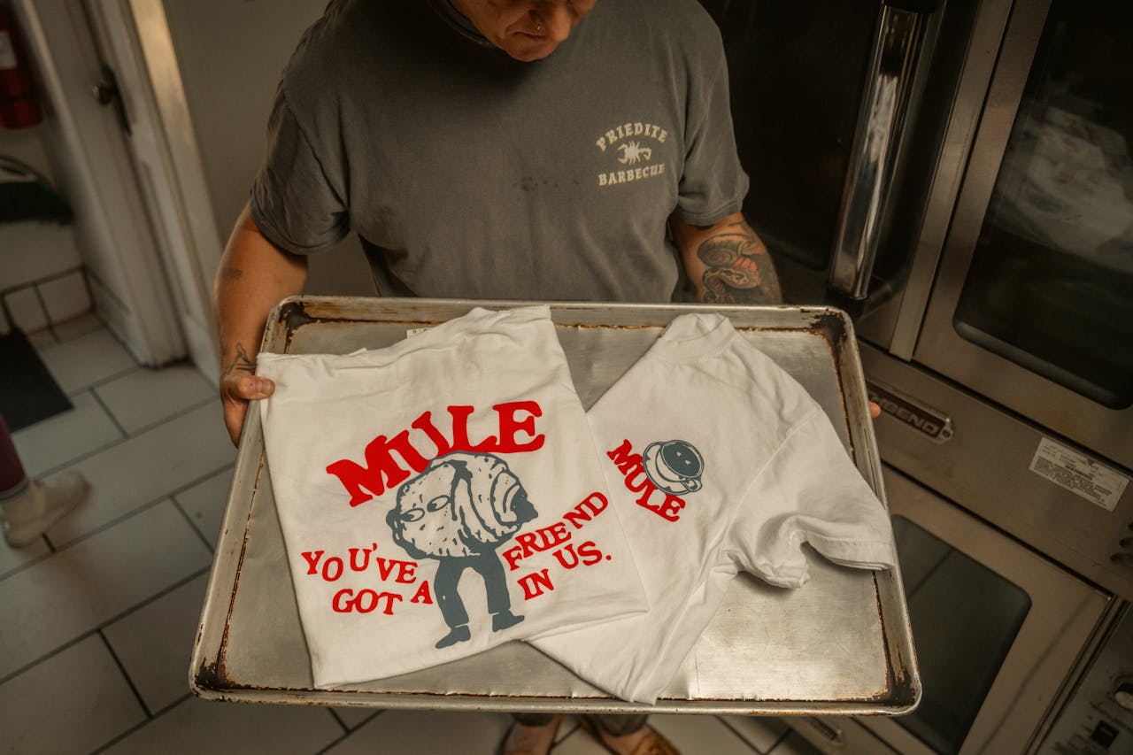 Person showcasing white T-shirts with red mule design and humorous text, indoors.